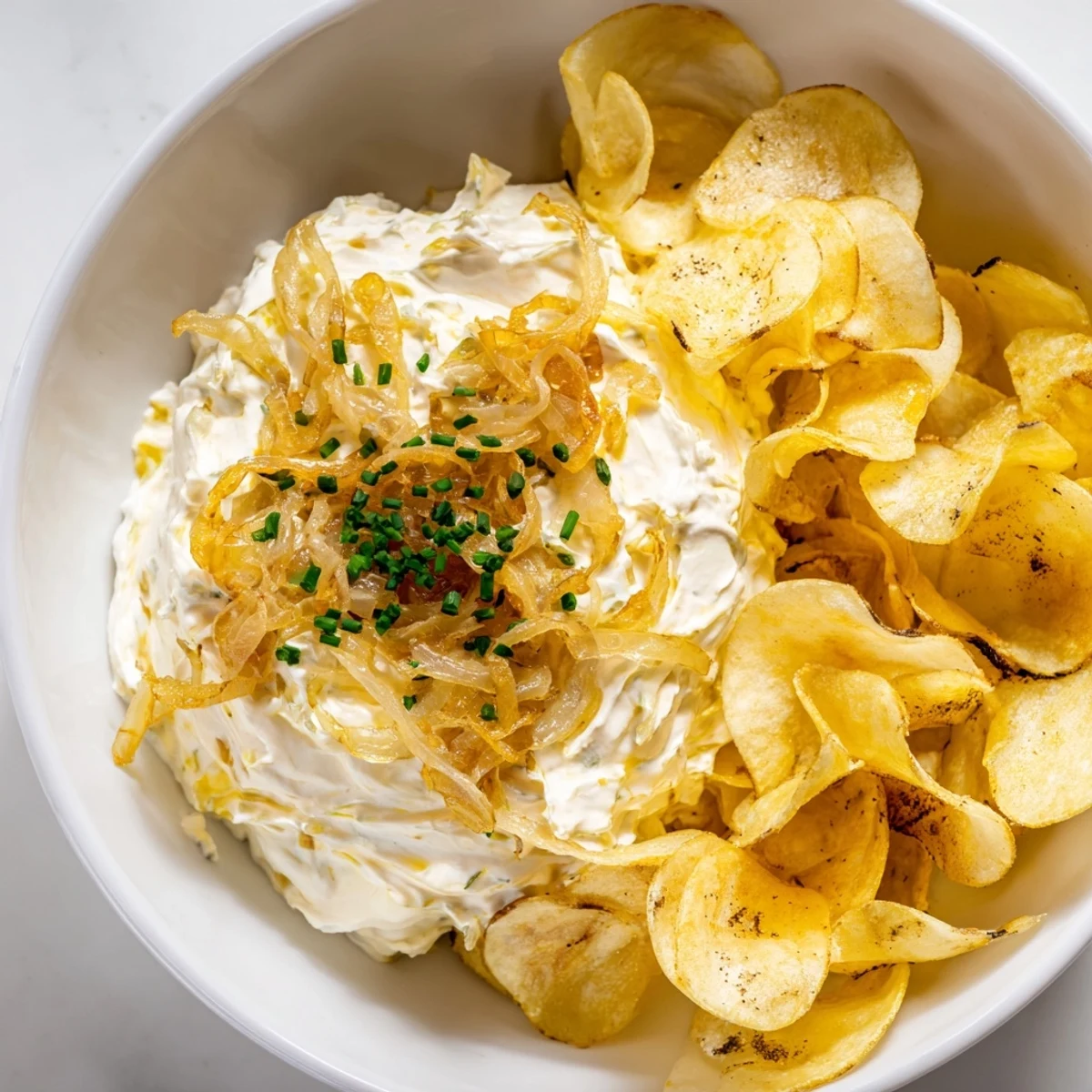 Freshly fried russet potato chips are stacked beside a rustic bowl of savory onion dip.