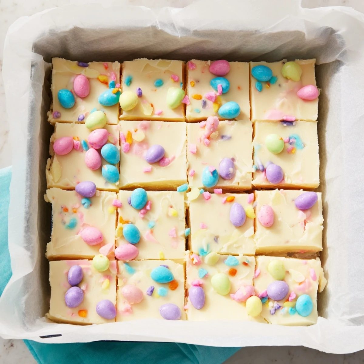 Creamy Easter Fudge squares topped with pastel candy eggs and sprinkles on a marble countertop.