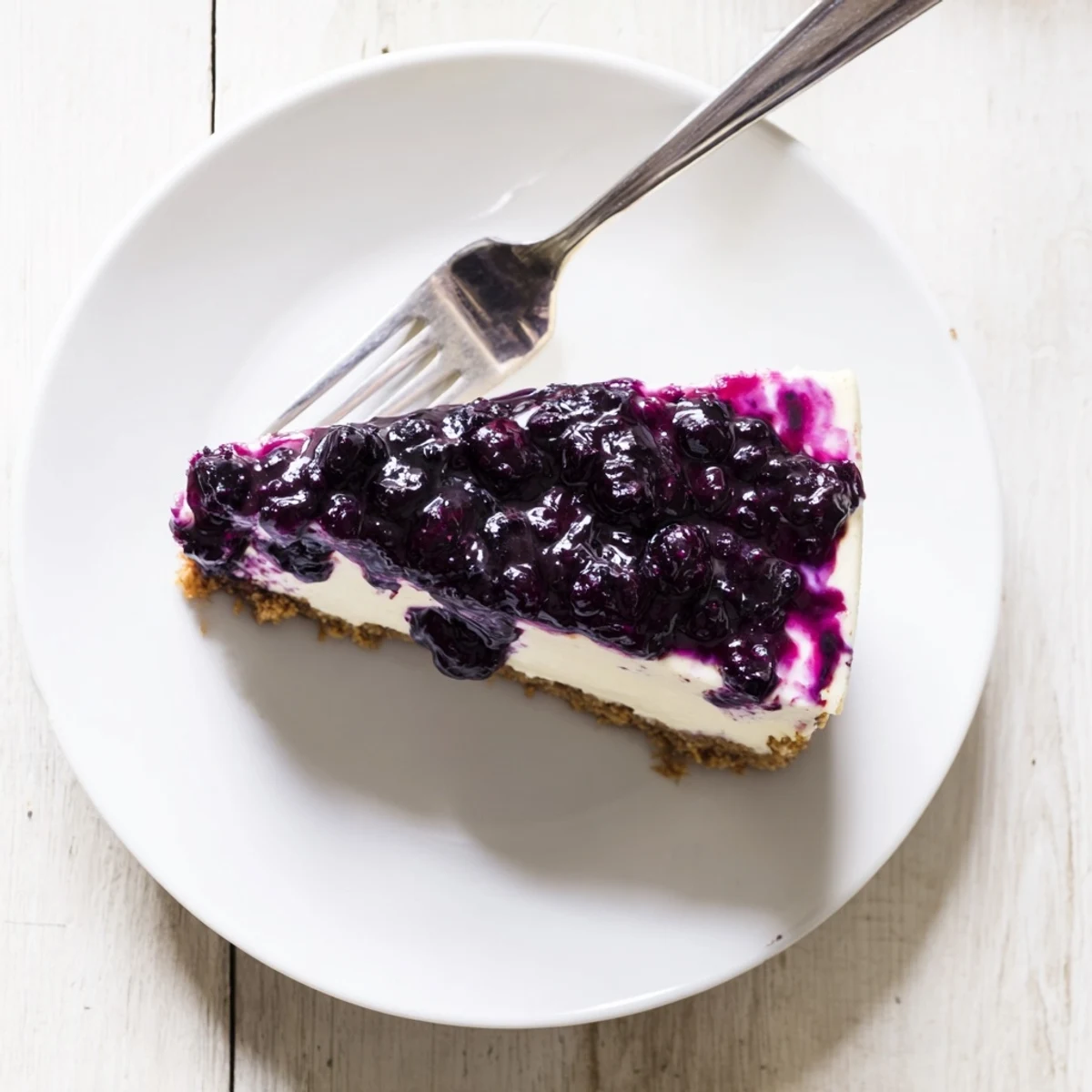 Creamy blueberry cheesecake slice showcasing velvety filling and graham cracker crust topped with glossy purple compote