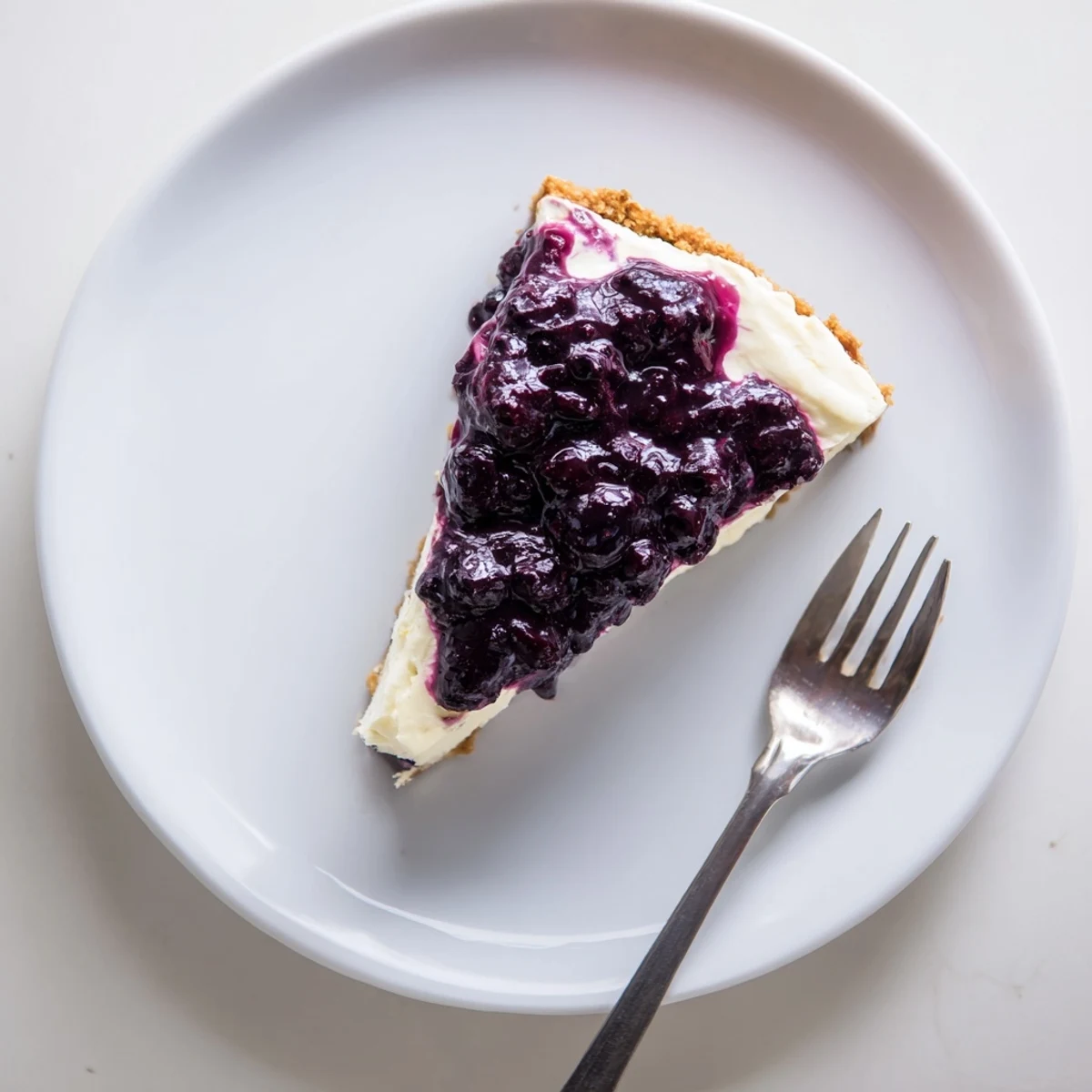 Indulgent blueberry cheesecake wedge revealing smooth cream cheese layer beneath sweetened fruit topping and crunchy crust