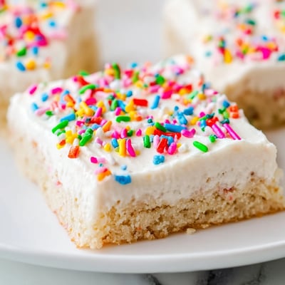 Freshly baked Sugar Cookie Bars stacked on a wire rack, showing their tender golden edges and smooth vanilla frosting.
